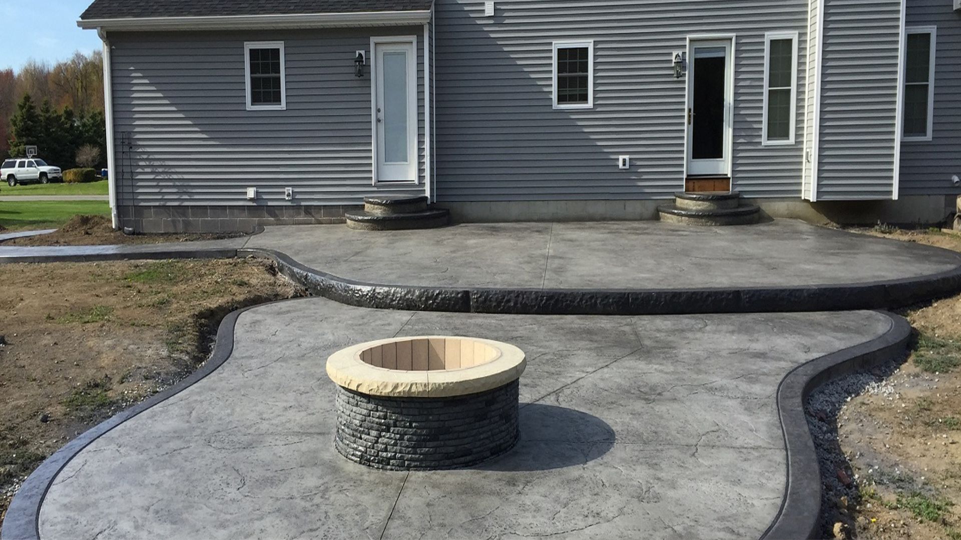 Curved concrete patio with stone fire pit near gray vinyl-sided house